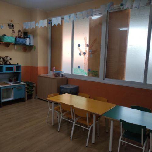 Aula con mesas rectangulares y sillas y zona de juegos en pared