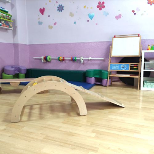 Aula grande con elementos de juego de madera, colchonetas y pizarra blanca al fondo