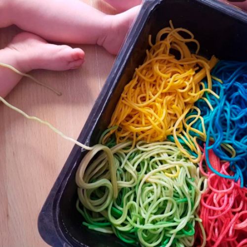 Piernas de bebé sentado junto a bandejas con espaguettis de colores